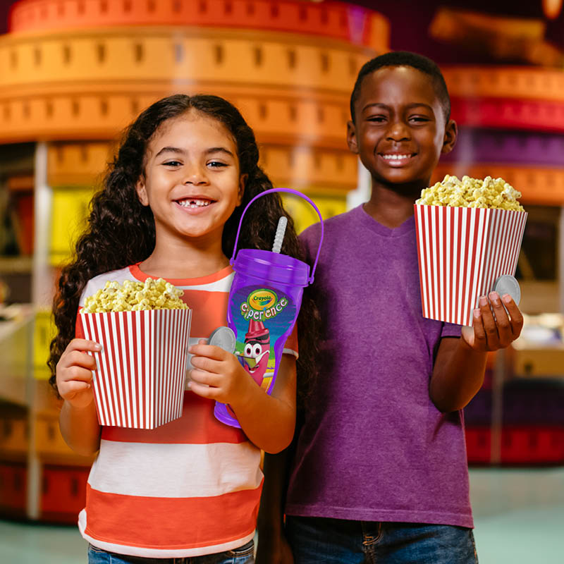Two kids holding out popcorn, a souvenir drink, and two tokens